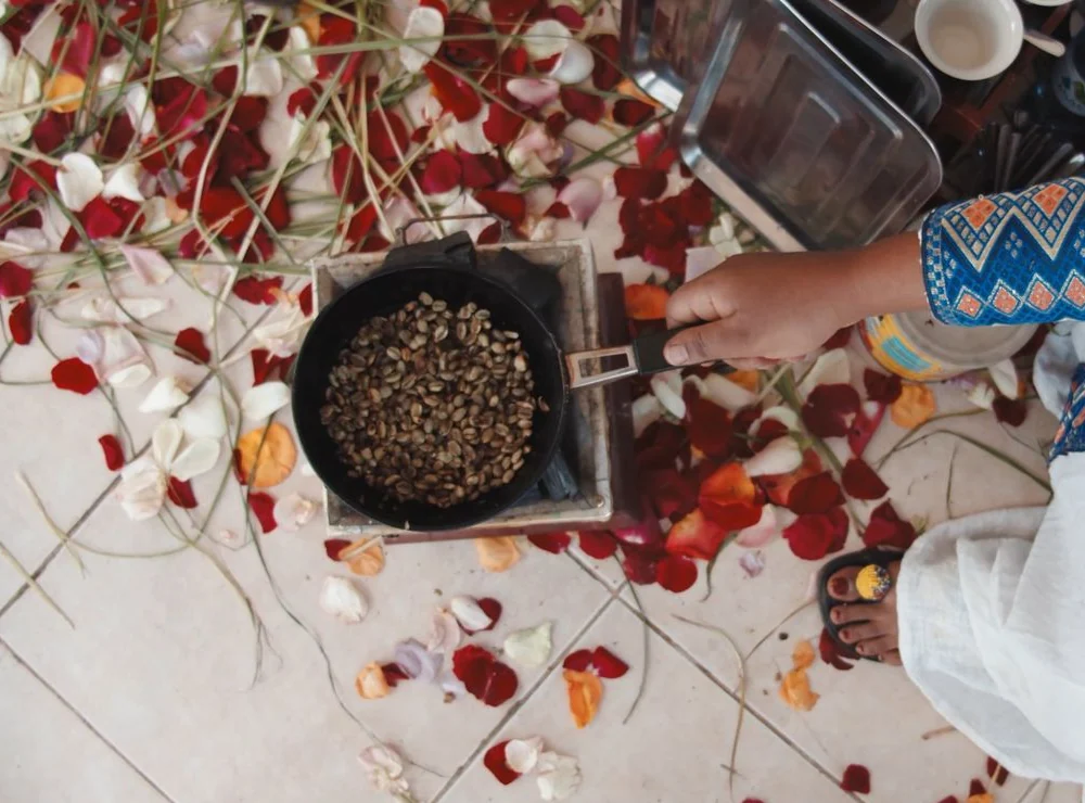 Coffee beans roasting in a pan over a brazier beside cups and flower petals